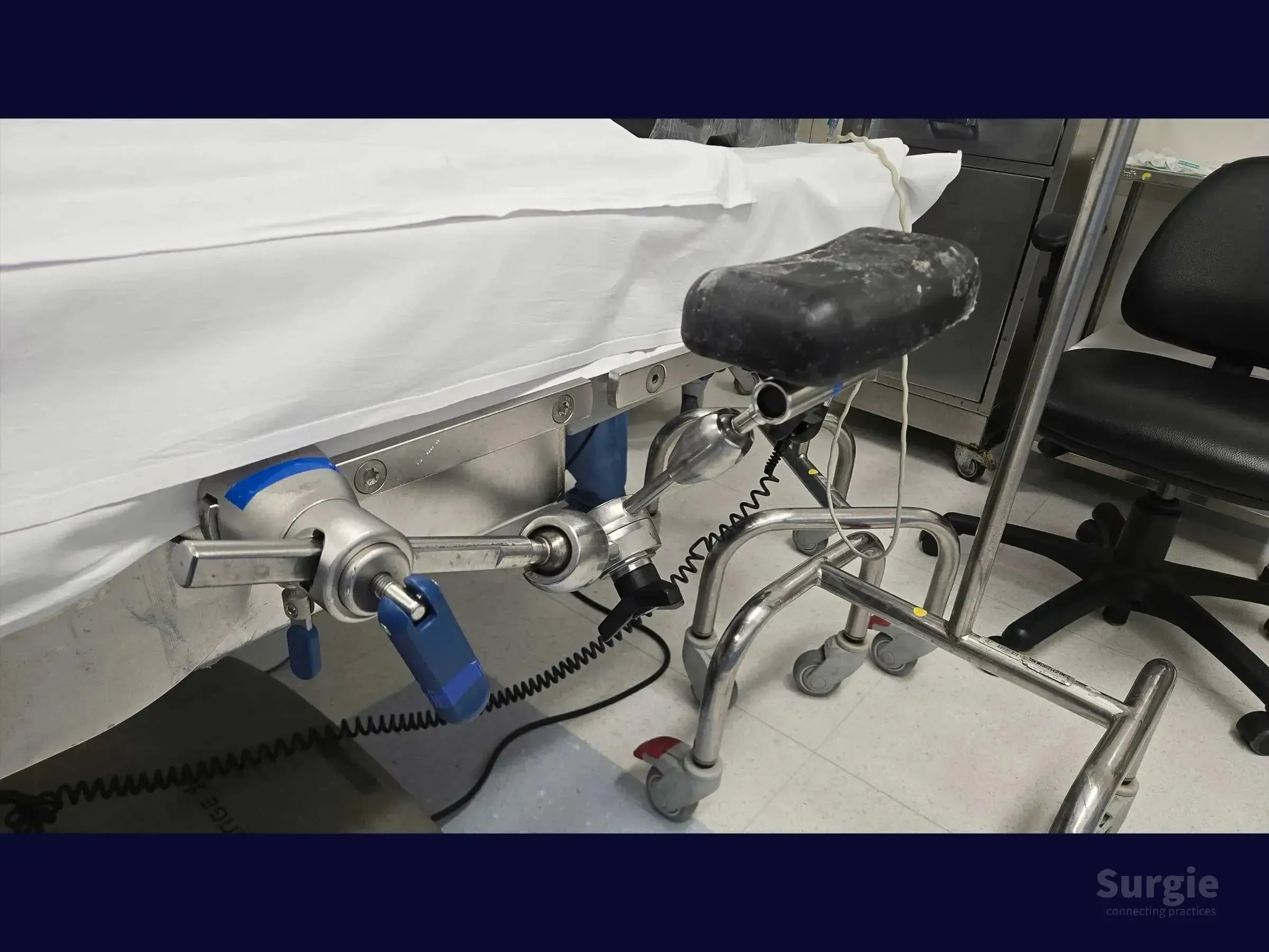 Surgical arm support attachment with adjustable metal joints and a black padded rest mounted to an operating table.